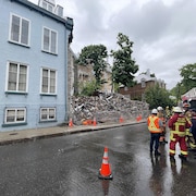 Une équipe de pompiers et des travaux publics regardent l'immeuble à gauche de celui qui a été démoli.