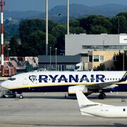 Un avion sur lequel on peut lire « Ryanair » est vu au sol près d'un autre avion. Un escalier permettant aux passagers de quitter l'avion est vu près de l'appareil.