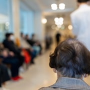 Le derrière de la tête d'une femme âgée dans une salle d'attente. Autour d'elle, on voit les contours flous de nombreuses personnes assises sur des chaises et d'un individu qui passe, vêtu d'un sarrau blanc.