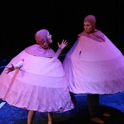 Sarah Bergbush et Bobbi Jones sur scène, habillées en seins géants. Elles portent un tissu rose pâle avec un mamelon sur leur tête. 