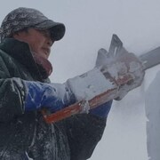 Gros plan sur un homme de profil habillé très chaudement et qui manie une scie pour façonner une sculpture de neige. Son manteau est couvert de neige et de la neige poudreuse revole dans son visage.
