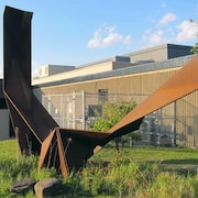 Une sculpture en acier repose sur un terrain près d’un bâtiment.