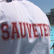 Un surveillant de baignade sur le bord d'une piscine extérieure publique. Le mot sauveteur est imprimé sur le dos de son chandail.