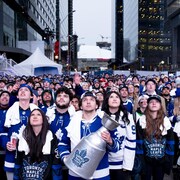 Des partisans des Maples Leafs rassemblés devant l'aréna banque Scotia, tous en ligne, les yeux rivés sur l'écran géant et portant le chandail de leur équipe, l'un d'entre eux tient une coupe Stanley à la main.