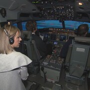On voit l'intérieur de la cabine du simulateur de vol. Un pilote et un co-pilote sont assis aux commandes (on les voit de dos). Derrière eux, l'instructrice parle.