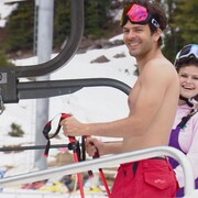 Un homme sans chandail avec des lunettes de ski sur la tête. Une femme est en arrière-plan. 