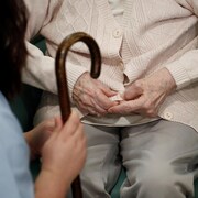 Une infirmière tient la canne en bois d'une personne âgée qui est assise face à elle et dont on ne voit que les mains.
