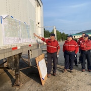 Un pompier forestier montre du doigt un point sur une carte affichée sur un conteneur, à l'extérieur. Cinq personnes le regardent. Tous portent l'uniforme de la Sopfeu: chemise rouge et pantalon bleu foncé. 