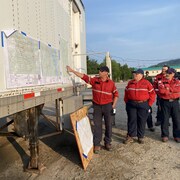 Un pompier forestier montre du doigt un point sur une carte affichée sur un conteneur, à l'extérieur. Cinq personnes le regardent. Tous portent l'uniforme de la Sopfeu: chemise rouge et pantalon bleu foncé. 