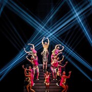 Sur une scène éclairée par des faisceaux lumineux, une femme chante sur des escaliers, entourée de danseurs et de danseuses.