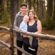 Une femme sourit en posant avec son jeune fils dans ses bras et son conjoint, derrière une clôture faite de branches d'arbre, devant un boisé.