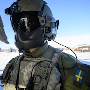 Un soldat masqué près d'un hélicoptère. Son uniforme porte un drapeau suédois.