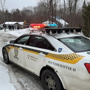 Une voiture de la Sûreté du Québec est stationnée dans une entrée de maison, entourée d'un boisé. Des gens de la SQ sont debout dans l'entrée. 