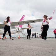 Des agentes de bord en chemises blanches et cravates roses sont sur le tarmac, sous une aile d'un avion peint en blanc et rose.