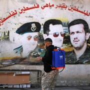 Un homme portant un masque médical désinfecter une rue devant un mur sur lequel on peut voir les portraits de l'ancien président Hafez Al-Assad et de ses deux enfants, Bachar, l'actuel président, et son frère Maher.