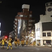 Un groupe de pompiers marchent près d'un immeuble sur le point de s'effondrer après un séisme. 