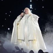 Un personnage vêtu d'une tunique blanche qui lui donne l'air d'un roi ancien chante sur scène.