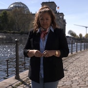 La journaliste Tamara Alt&eacute;resco est debout, sur un trottoir pav&eacute; de Berlin. Elle se trouve pr&egrave;s de l'eau. Derri&egrave;re elle, on voit le parlement allemand.