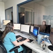 Une dame assise devant plusieurs écrans d'ordinateurs. derrière les écrans se trouve une vitre qui donne sur la chambre des patients. 