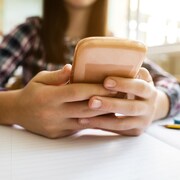 Un gros plan sur les mains d'une jeune fille qui tient un téléphone intelligent. Ses mains sont posées sur son pupitre d'école, par dessus un cahier ligné.