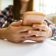 Un gros plan sur les mains d'une jeune fille qui tient un téléphone intelligent. Ses mains sont posées sur son pupitre d'école, par-dessus un cahier ligné.