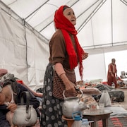 Une vieille femme, debout sous une immense tente qui abrite plusieurs personnes, tient une bouilloire dans sa main.