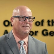 Un homme avec des lunettes vêtu d'un complet gris devant un mur jaune.