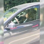 Capture d'écran d'une vidéo, une Tesla grise vue de côté, fenêtre du conducteur en gros plan. Au volant, une personne dont le visage est flouté est inclinée vers la droite, la tête penchée, une main posée sur le volant.