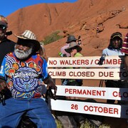 Des Aborigènes sont assis devant une montagne rouge et des inscriptions qui indiquent que la randonnée est interdite.