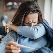 Une femme malade tousse dans son coude, un thermomètre à la main.