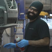 Un homme sourit dans un garage devant une voiture surélevée.
