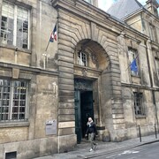 Un drapeau européen et un drapeau français sont disposés chaque côté de l'entrée du tribunal dans un immeuble historique.