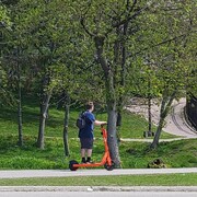 Un jeune homme, sac au dos, roule en trotinette électrique sur le trottoir, près d'un parc.