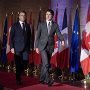Emmanuel Macron et Justin Trudeau marchent souriant devant une rangée de drapeaux. 