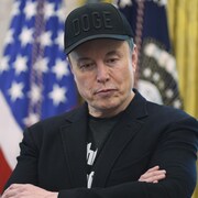 Devant un drapeau américain, Elon Musk, coiffé d'une casquette noire arborant le mot «DOGE», regarde vers le bas.