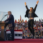 Donald Trump, les yeux fermés, parle au micro tandis qu'Elon Musk, les bras dans les airs, saute à ses côtés sur la scène d'un rassemblement extérieur.