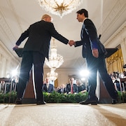 Donald Trump et Justin Trudeau se serrent la main devant les caméras et un groupe de personnes.