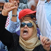 Dans une foule compacte, une femme voilée et d'autres manifestants crient en levant un bras dans les airs.