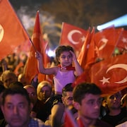 Des centaines de personnes brandissent des drapeaux de la Turquie, dont une petite fille qui est sur les épaules de son père. 