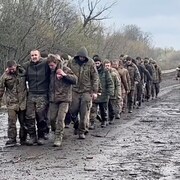 Une colonne de soldats marche sur une route, les premiers portant un homme visiblement blessé.