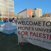 Des tentes sont plantées sur le campus de l'université. Sur une banderole on peut lire « Bienvenue à l'Université du peuple pour la Palestine ». Puis, « pour le bien public ».