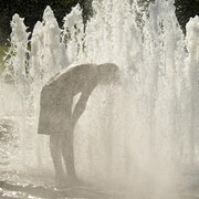 Une personne accablée par la chaleur se tient penchée au-dessus du jet d'une fontaine pour se rafraîchir.
