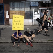 Deux personnes sont assises sur le trottoir, dont une femme qui tient une pancarte sur la décriminalisation.