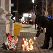 Une personne allume des lampions près de peluches déposées devant une église, le soir, en hiver. 