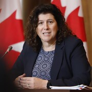 La vérificatrice générale du Canada Karen Hogan parle au micro assise à une table devant des drapeaux canadiens.