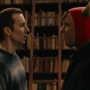 Deux hommes, dont l'un est déguisé en démon, se font face devant une bibliothèque remplie de livres.