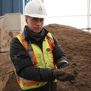 Un homme avec une veste jaune fluorescente et un casque blanc tient du compost dans ses mains.