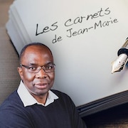 Jean-Marie Yambayamba sourit devant l'image d'un livre dans lequel est écrit «Les carnets de Jean-Marie» et sur lequel est posée une plume fontaine.