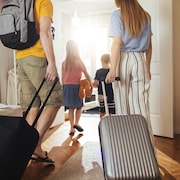 Deux adultes et deux enfants photographiés de dos, sortant de leur domicile. Les deux adultes tirent des valises.