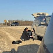 Le premier ministre, en costume-cravate, qui s'active à changer un pneu sur le bord de la route.